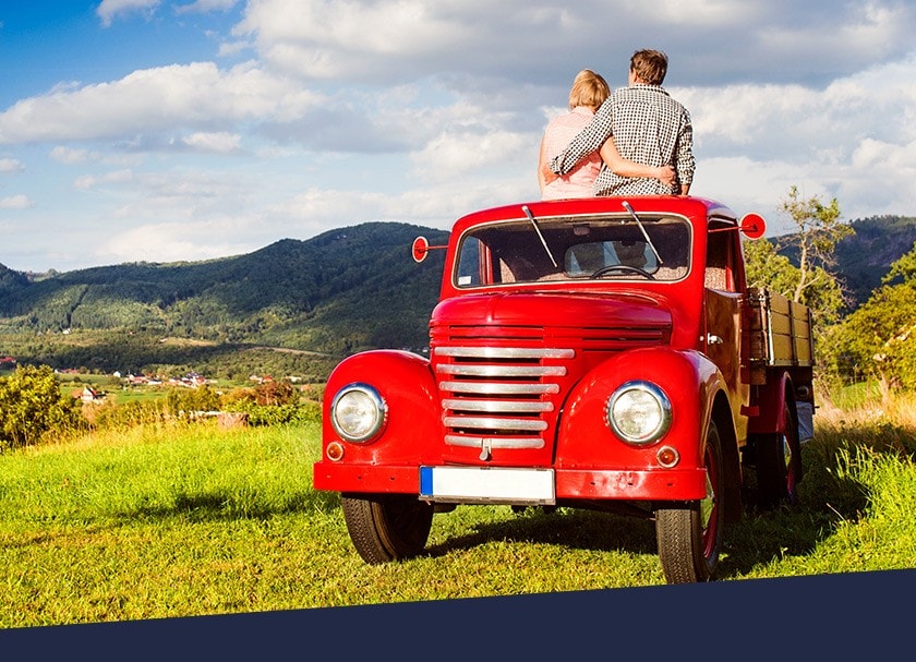 A couple sitting on the roof of a red truck in a farm field admire the landscape