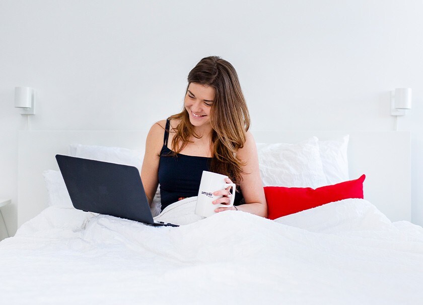 A woman uses her laptop in bed while holding a Simplii mug