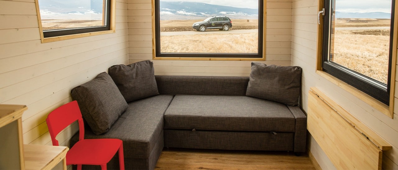 A tiny home with pull-out sofa, folding table and large windows showing mountains and a car